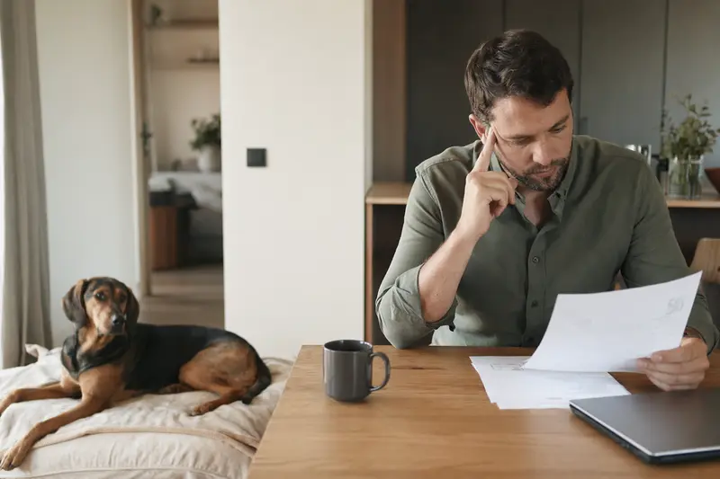 Ein Hundebesitzer sitzt mit einer Tierarztrechnung am Tisch, während ein entspannter Pannonischer Spürhund ruhig auf einer Decke liegt.