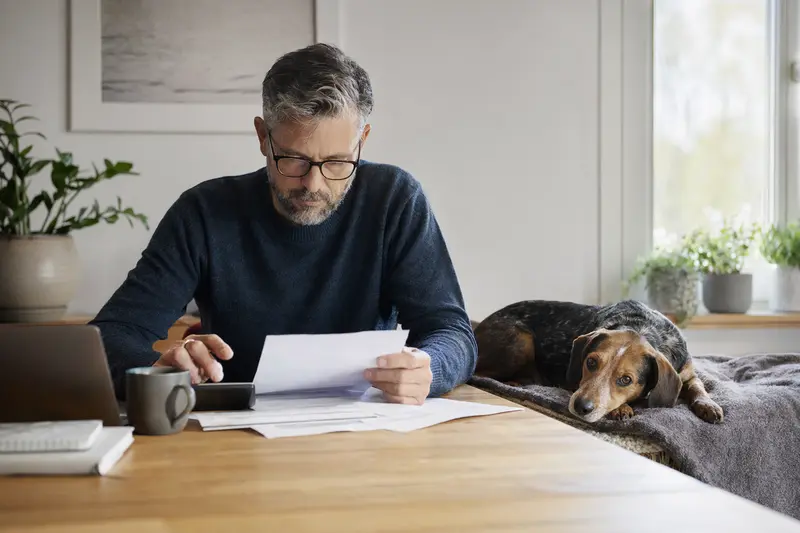 Ein Hundehalter sitzt mit einer Tierarztrechnung am Tisch, während eine entspannte Polnische Bracke auf einer Decke liegt und die Situation ruhig bleibt.