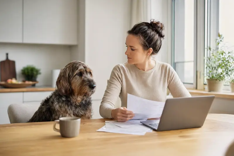 Ein Polnischer Niederungshund sitzt ruhig neben seinem Besitzer am Küchentisch, während Versicherungsunterlagen und ein Laptop mit Vergleichsseite auf eine Hundeversicherung hinweisen.