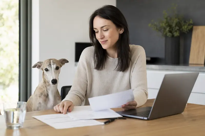 Ein Polnischer Windhund sitzt ruhig neben seinem Besitzer am Küchentisch mit Versicherungsunterlagen und Laptop für eine Hundeversicherung.