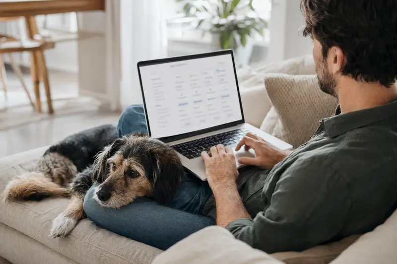 Ein Hundebesitzer sitzt mit einem Laptop auf dem Sofa, während ein treuer Hund daneben entspannend liegt.