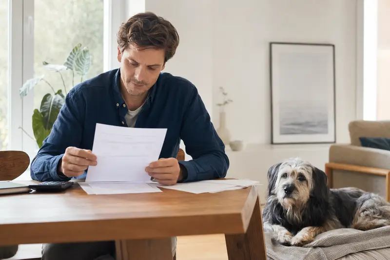 Ein Hundebesitzer prüft eine Tierarztrechnung am Tisch, während ein entspannter Pyrenäenschäferhund auf einer Decke daneben liegt.