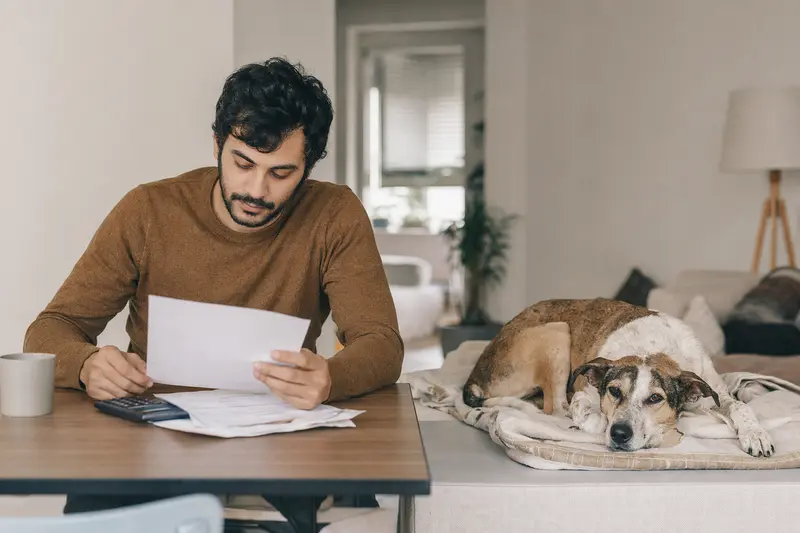 Ein Hundehalter sitzt mit einer Tierarztrechnung am Tisch, während ein entspannter Rampuri Hund auf einer Decke daneben liegt und das Thema Hundekrankenversicherung ruhig und realistisch vermittelt.