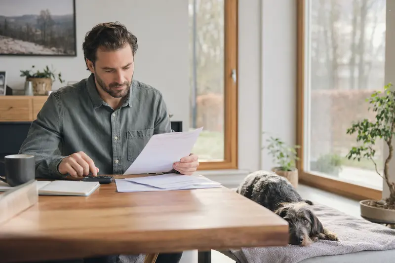Ein Hundehalter sitzt mit einer Tierarztrechnung am Tisch, während seine Rauhhaarige Istrianer Bracke entspannt auf einer Decke liegt, passend zum Thema Hundeversicherung.