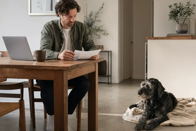Ein Hundebesitzer sitzt mit einer Tierarztrechnung am Tisch, während ein entspannter Riesenschnauzer ruhig auf einer Decke liegt – ein passendes Motiv zu Tierarztkosten und Hundeversicherung.