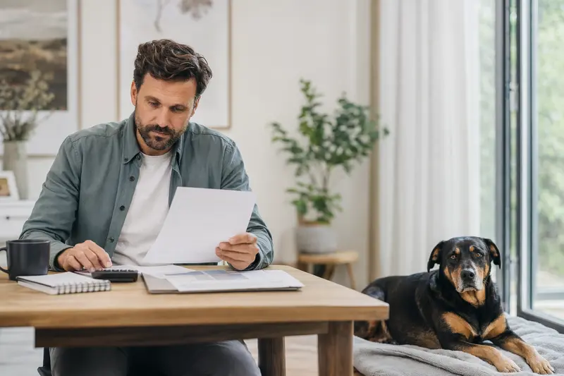 Ein Hundehalter sitzt mit einer Tierarztrechnung am Tisch, während ein entspannter Rottweiler auf einer Decke liegt – ein ruhiges Bild zum Thema Hundekrankenversicherung und Tierarztkosten.