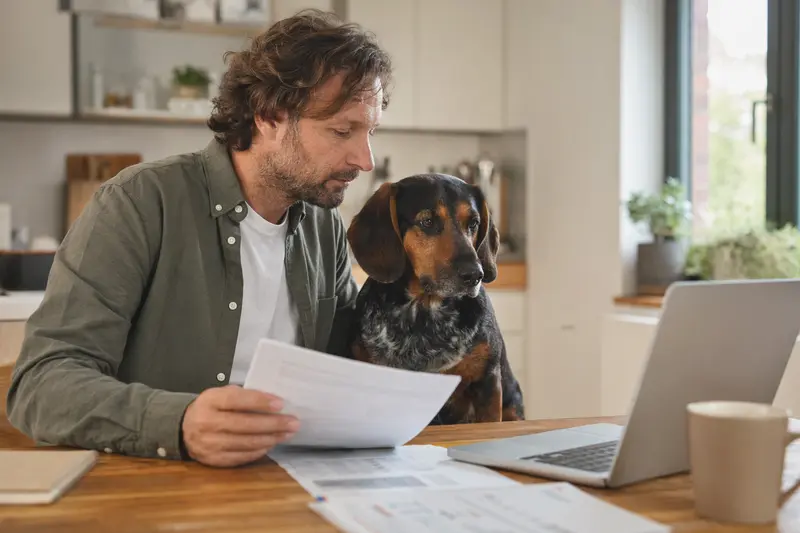 Eine Sauerländer Dachsbracke sitzt ruhig neben ihrem Besitzer am Küchentisch, während Versicherungsunterlagen und ein Laptop mit einer Vergleichsseite auf dem Tisch liegen.