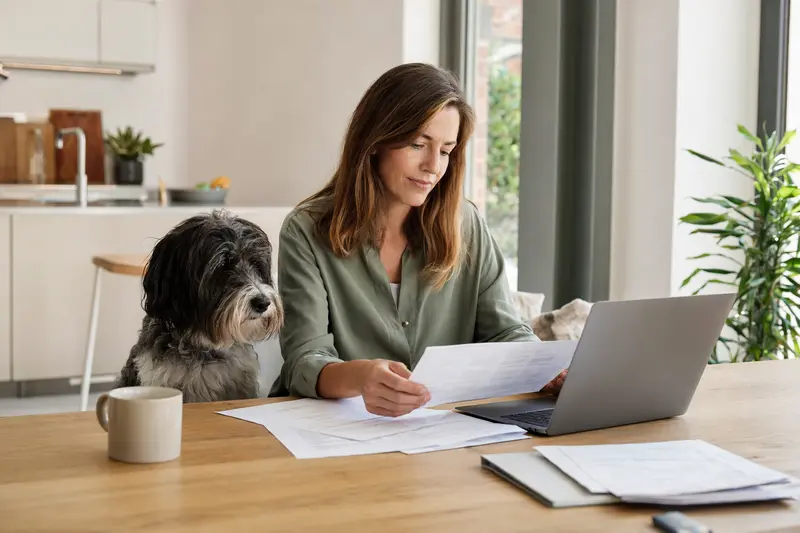 Ein Schapendoes sitzt ruhig neben seinem Besitzer am Küchentisch, während Versicherungsunterlagen und ein Laptop mit einer neutralen Vergleichsseite auf eine Hundeversicherung hinweisen.