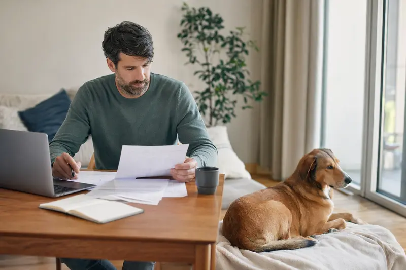 Ein Hundebesitzer sitzt mit einer Tierarztrechnung am Tisch, während sein entspannter Schiller-Laufhund auf einer Decke liegt und der Moment ruhig und lösungsorientiert wirkt.