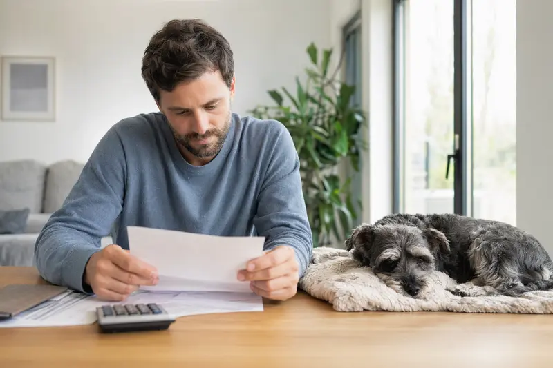 Ein Hundebesitzer sitzt mit einer Tierarztrechnung am Tisch, während ein entspannter Schnauzer auf einer Decke liegt – ein ruhiges Motiv zum Thema Tierarztkosten und Absicherung für Hunde.