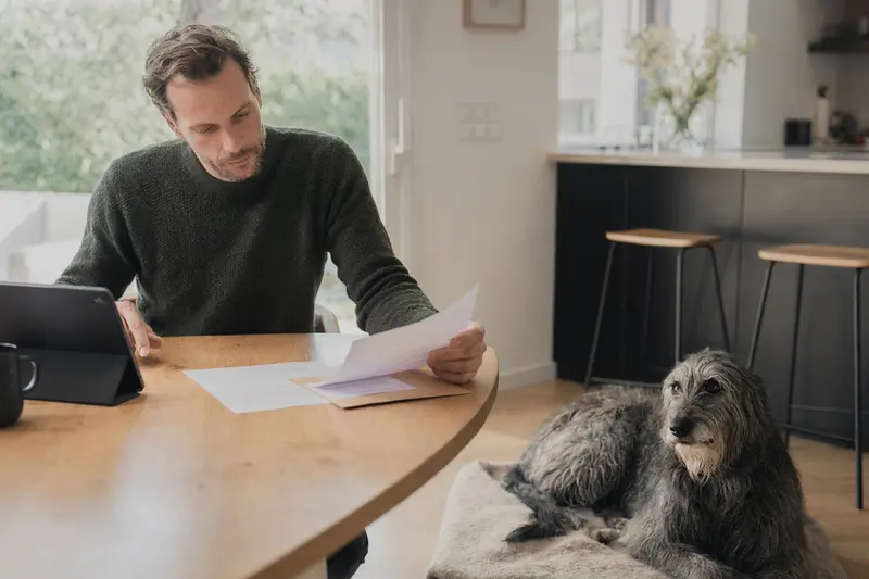 Ein Hundehalter sitzt mit einer Tierarztrechnung am Tisch, während ein entspannter Schottischer Hirschhund auf einer Decke neben ihm liegt.