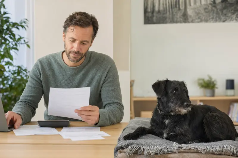 Besitzer sitzt mit Tierarztrechnung am Tisch, während ein entspannter schwarzer Terrier auf einer Decke liegt und die Haustierkrankenversicherung im Alltag veranschaulicht.