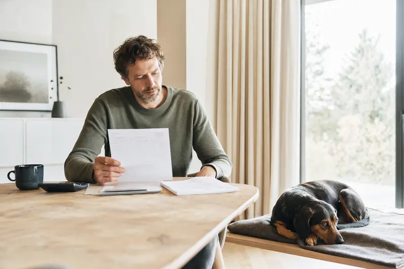 Ein Hundehalter sitzt mit einer Tierarztrechnung am Tisch, während ein entspannter Schwarzwildbracke-Hund auf einer Decke neben ihm liegt.