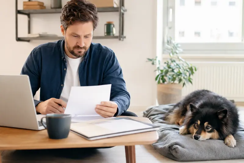 Ein Hundebesitzer sitzt mit einer Tierarztrechnung am Tisch, während ein entspannter Schwedischer Lapphund auf einer Decke daneben liegt.