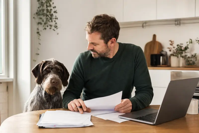Ein Slowakischer Rauhbart sitzt ruhig neben seinem Besitzer am Küchentisch mit Versicherungsunterlagen und einem Laptop zur Hundekrankenversicherung.