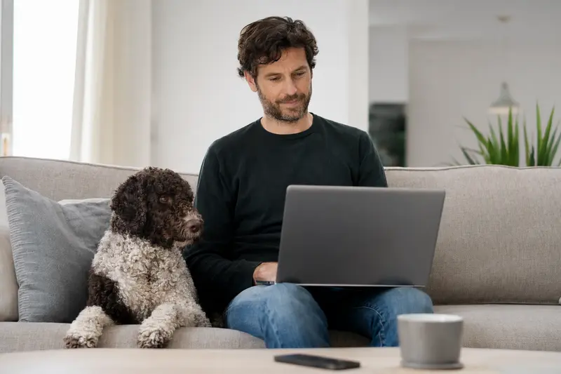 Ein Hundebesitzer sitzt mit seinem Spanischen Wasserhund auf dem Sofa und vergleicht Versicherungen auf einem Laptop.
