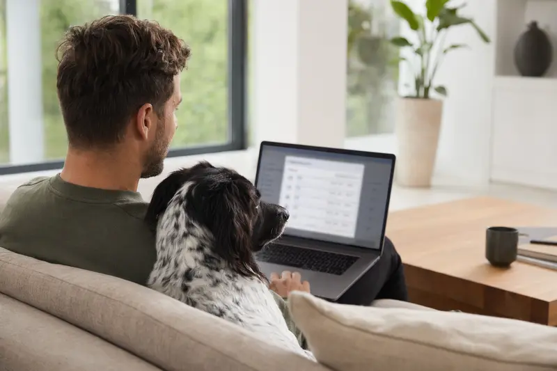 Ein Hund liegt neben seinem Besitzer auf dem Sofa, während dieser am Laptop einen Versicherungsvergleich für Haustiere durchführt.