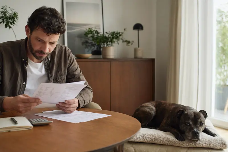 Ein Hundehalter sitzt mit einer Tierarztrechnung am Tisch, während ein entspannter Staffordshire Bullterrier auf einer Decke daneben liegt.