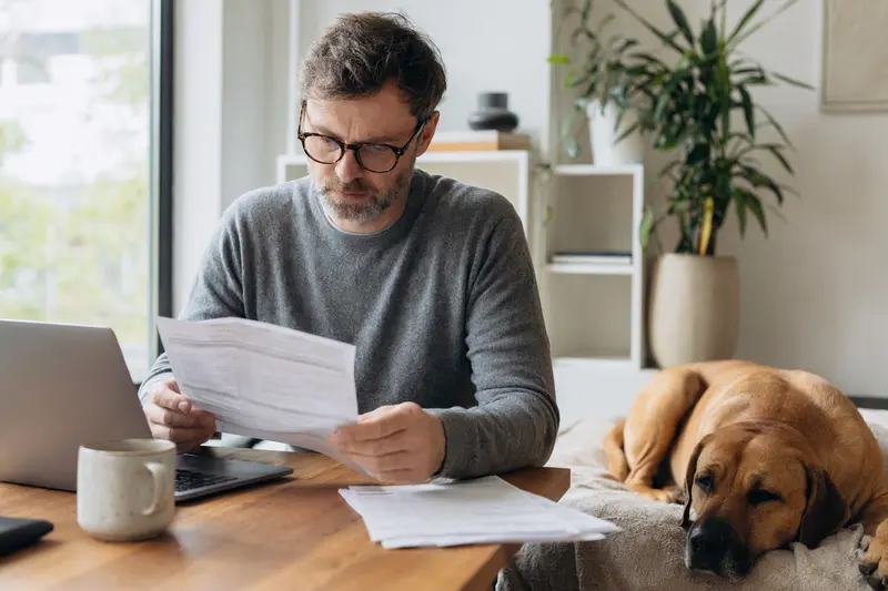 Ein Hundebesitzer sitzt mit einer Tierarztrechnung am Tisch, während ein entspannter Tosa Inu auf einer Decke liegt und das Thema Tierarztkosten für Hunde ruhig und seriös vermittelt.