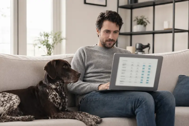 Ein Hund neben seinem Besitzer, der auf dem Laptop in einer ruhigen Wohnung nach Tierversicherungen sucht.