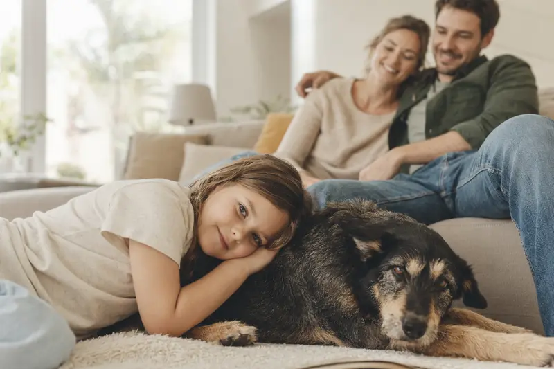 Ein freundlicher Hund liegt entspannt neben einem Kind im hellen Wohnzimmer, während die Eltern im Hintergrund eine sichere Familienatmosphäre schaffen.