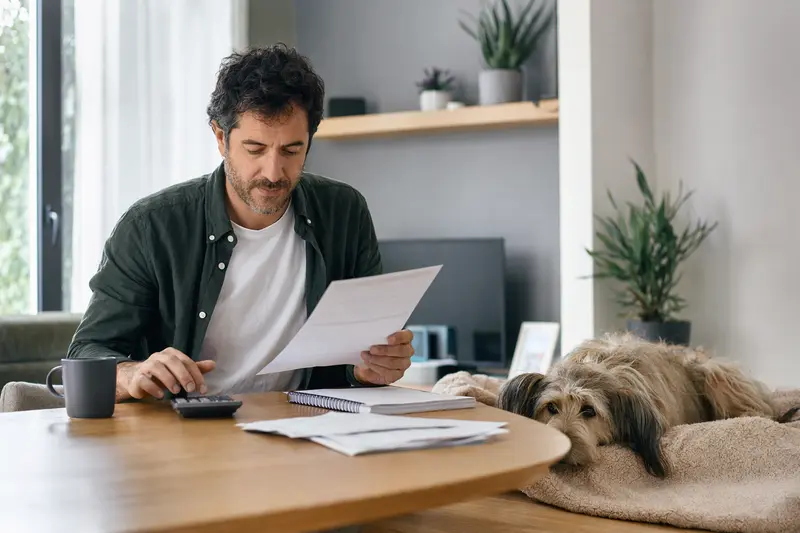 Ein entspannter Villanuco de Las Encartaciones Hund liegt neben einem Hundebesitzer mit einer Tierarztrechnung am Tisch in einer hellen Wohnung.