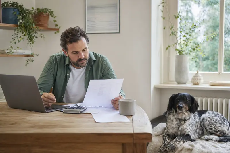 Ein Hundehalter sitzt mit einer Tierarztrechnung am Tisch, während ein entspannter Westerwälder Kuhhund ruhig auf einer Decke liegt.
