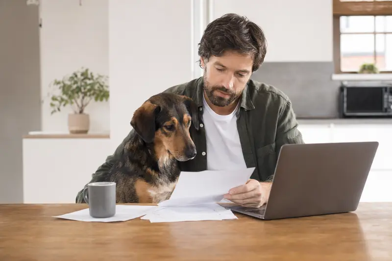 Ein Westerwälder Kuhhund sitzt ruhig neben seinem Besitzer am Küchentisch, während Versicherungsunterlagen und ein Laptop mit einer neutralen Vergleichsseite liegen.