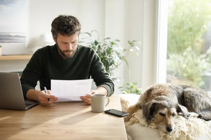 Ein Hundehalter sitzt mit einer Tierarztrechnung am Tisch, während ein entspannter Hund auf einer Decke daneben liegt und die ruhige Wohnatmosphäre eine lösungsorientierte Situation rund um Tierarztkosten vermittelt.